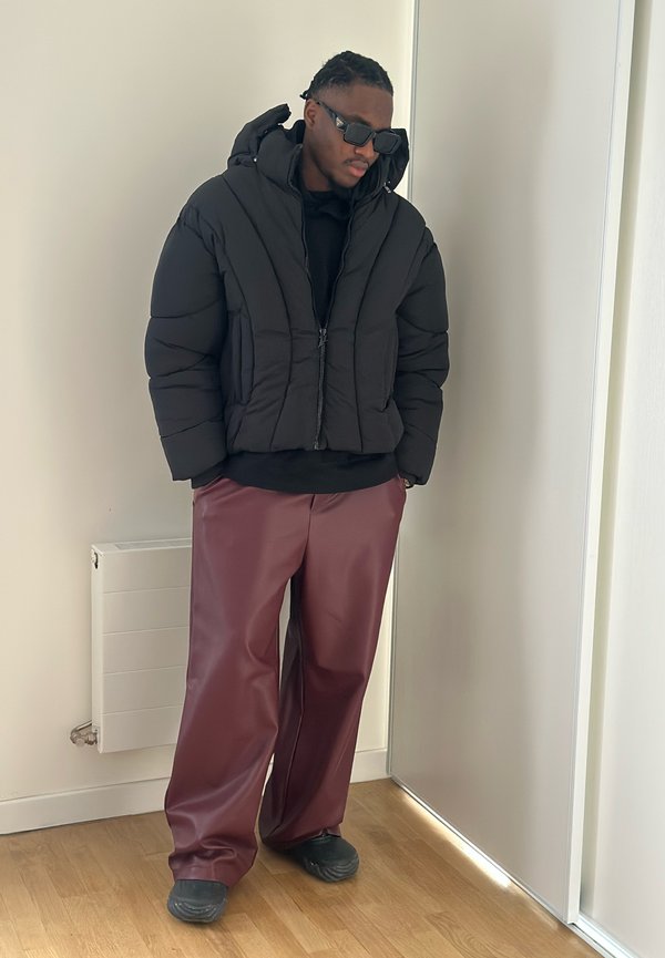 Moški oblečen v črno puhasto jakno, črna sončna očala, burgundske široke hlače in črni čevlji stoji v notranjosti na lesenem tleh.