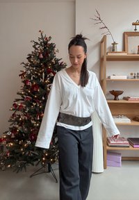 Haut blanc à manches longues avec un col en V, associé  à un pantalon noir et une ceinture texturée. En arrière-plan, un sapin de Noël décoré.