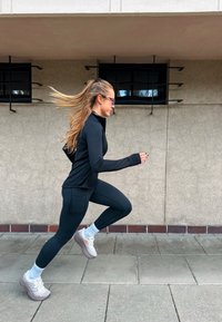 Nais is Blacki treeningvarustuses ja valgetes tossudes jookseb väljas sillutatud pinnal, tema taga on beež sein ja restiga aknad.