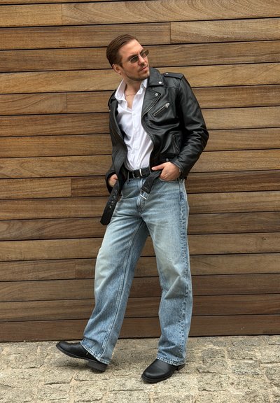 Hombre con chaqueta de cuero negra, camisa blanca, vaqueros azul claro y botas negras posa contra una pared de madera con las manos en los bolsillos.