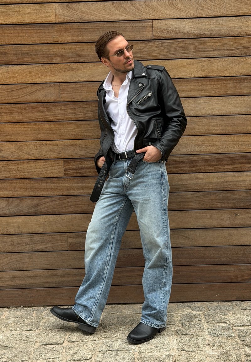 Homme portant une veste en cuir noire, une chemise blanche, un jean bleu clair et des bottes noires pose contre un mur en bois, les mains dans les poches.