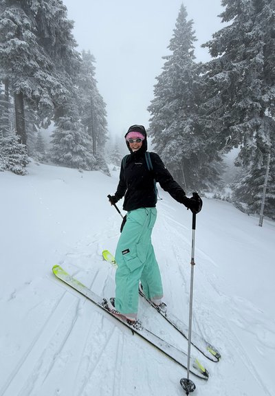 Persona con chaqueta negra y pantalones verde menta esquiando en un sendero cubierto de nieve rodeado de pinos helados en un día neblinoso.
