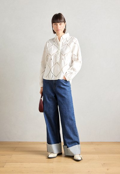 Blusa blanca con estampado de manga larga y escote con botones, combinada con unos vaqueros de mezclilla azul anchos con dobladillo deshilachado y puños en contraste.