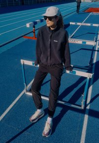 Sportaš sjedi na prečki za prepreke, odjeven u crnu flis jaknu, crne hlače i višebojne tenisice za trčanje, s laganom kapom i sunčanim naočalama.