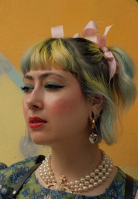 Πράσινη μπλούζα με λουλουδάτο μοτίβο, στρωτή κοσμηματοθήκη με πέρλες, χρυσά σκουλαρίκια σε σχήμα καρδιάς, ανοιχτόμπλε και κίτρινα μαλλιά, ροζ κορδέλες στα μαλλιά.