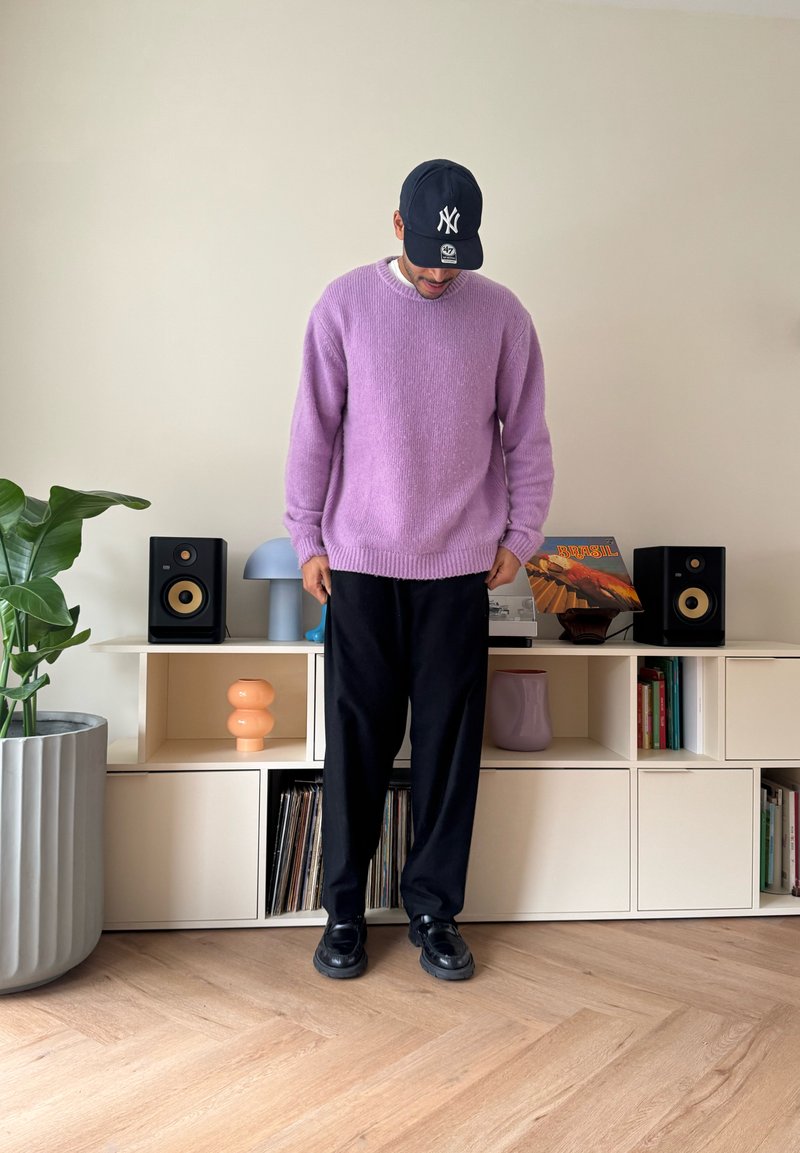 Homme portant un pull violet, un pantalon noir et une casquette noire, debout à l'intérieur près d'une étagère avec des haut-parleurs, un tourne-disque et des disques vinyles.