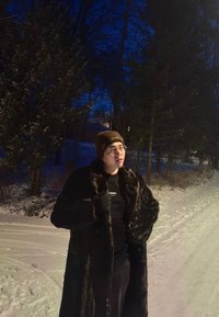 Personne portant un manteau en fourrure sombre, un bonnet marron et des lunettes, se tenant sur un chemin enneigé au crépuscule, avec des arbres et une maison en arrière-plan.