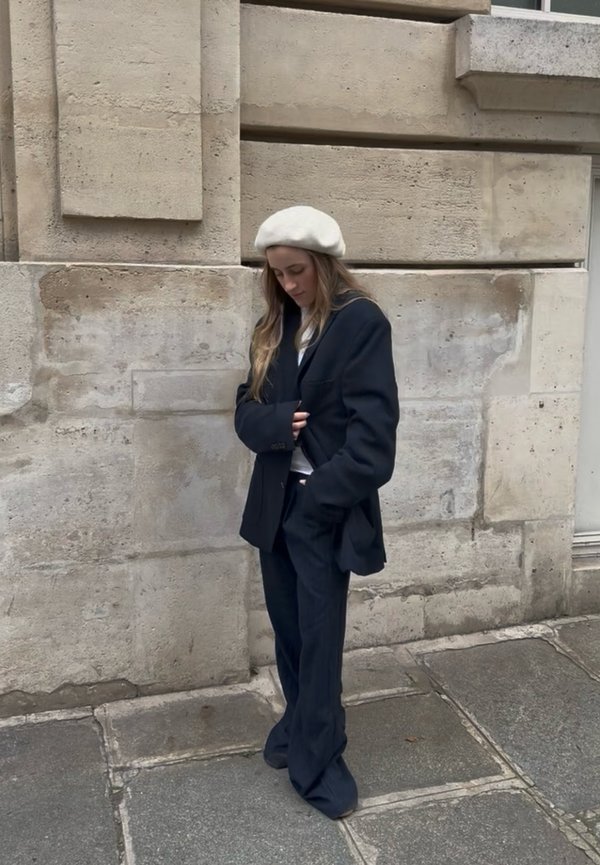 Žena v bielom barete a nadrozmernom čiernom obleku stojaca na kamennom chodníku pri béžovej kamennej stene, pozerá sa dole.