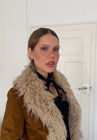 Una giovane donna con capelli intrecciati indossa grandi orecchini circolari, una giacca marrone con colletto di pelliccia beige e una sciarpa nera annodata intorno al collo.