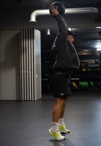 Črno fitnes žogo drži nad glavo oseba, oblečena v sivo kapuco, črne kratke hlače in bele športne copate z neon rumenimi detajli v telovadnici.