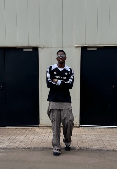 Camiseta de jersey de algodón negra con rayas blancas, combinada con pantalones cargo grises. De pie frente a puertas oscuras, usando gafas de sol y zapatos negros.