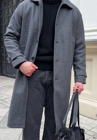 Cappotto grigio di lana con collo a risvolto, chiusura con bottoni e tasche laterali. Indossato sopra un maglione nero e abbinato a jeans scuri, tiene una borsa.