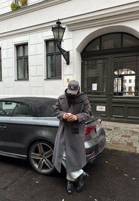 Sivi volneni plašč z ujemajočo se rutko, črni usnjeni škornji, bele hlače, siva kapa z logotipom, naslonjen na temno sivi kabriolet.