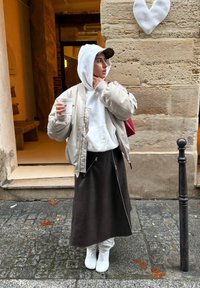 Femme en veste beige et sweat à capuche blanc tenant une tasse de café, debout dans une rue pavée à côté d'un mur en pierre avec une décoration en forme de cœur blanc.