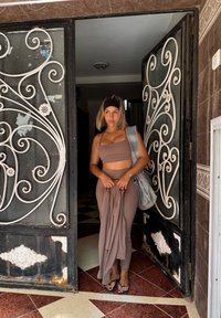 Jonge vrouw in bruine sportkleding en hoofdband staat in een deuropening met sierlijke zwart-witte smeedijzeren poorten, terwijl ze een jas en een denim tas vasthoudt.