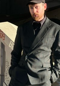 Costume noir à fines rayures avec un tissu texturé, présentant un revers cranté et un bouton unique. Accompagné d'une chemise boutonnée sombre et d'une casquette.