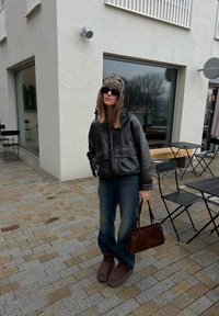 Femme portant une veste en cuir noire, un chapeau en fourrure, des lunettes de soleil, un jean bleu et des bottes marron, tenant un sac à main marron sur une terrasse de café.
