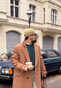 Manteau en laine couleur fauve par-dessus un pull vert, tenant une tasse de café dans un porte-gobelet en carton, avec un sac marron, marchant devant une voiture vintage bleue.