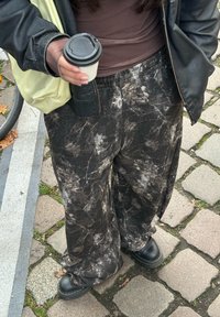 Pantalon large marbré noir et blanc, tissu doux, associé à des chaussures épaisses noires. Un sac léger et une tasse de café tenue à la main.