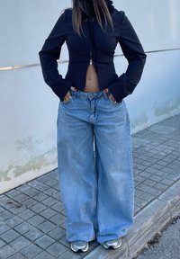 Črna prilegajoča jakna z zadrgo in kapuco, kombinirana s širokimi modrimi denim hlačami. Obuta v superge.