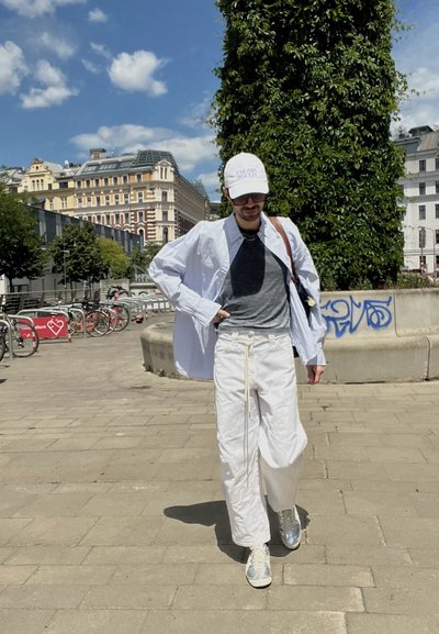 Camisa blanca de gran tamaño, camiseta gris, pantalones anchos blancos con cordón, zapatillas plateadas y una gorra blanca. El fondo muestra arquitectura urbana.