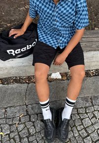 Osoba sjedi na drvenoj klupi, nosi crne kratke hlače, plavu košulju s kariranim uzorkom, bijele Puma čarape, crne cipele, drži crnu Reebok torbu.