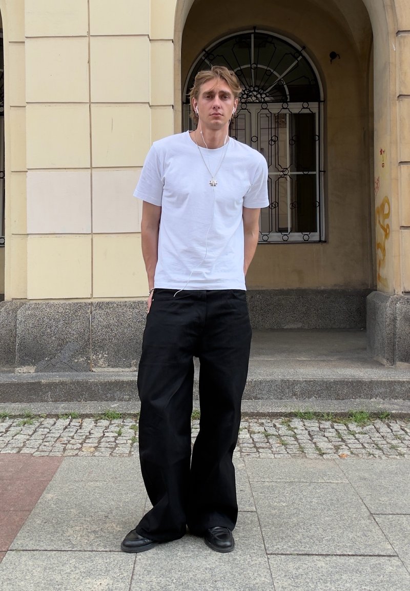 Jeune homme avec des écouteurs portant un t-shirt blanc et un pantalon large noir, debout sur un pavé en pierre près de l'entrée d'un bâtiment.