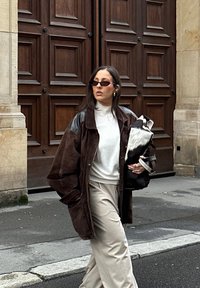Oversized brun ruskindjakke med sort læderærmer, hvid højhalset trøje, beige bukser og en sort-hvid tekstureret håndtaske.