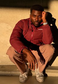 Μαρόν φουσκωτό μπουφάν με φερμουάρ μπροστά, μπεζ παντελόνι, λευκά αθλητικά παπούτσια και ασημένιες βέρας. Το υποκείμενο είναι κουλουριασμένο μπροστά σε μια υφή φόντου.