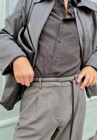 Sivi odijelo s prugicama uz crnu zip-jaknu i tamno prugastu košulju. Crna torba. Vidljivi detalji uključuju gumbe i šnalu na hlačama.