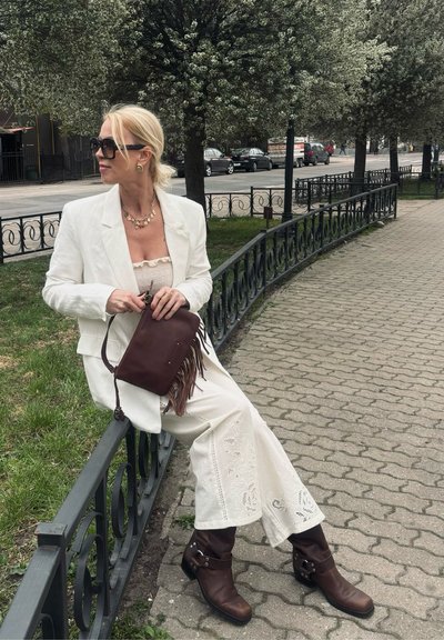 Mujer con traje blanco y botas marrones sentada en una verja metálica negra junto a la acera, sosteniendo un bolso marrón con flecos bajo árboles en flor.