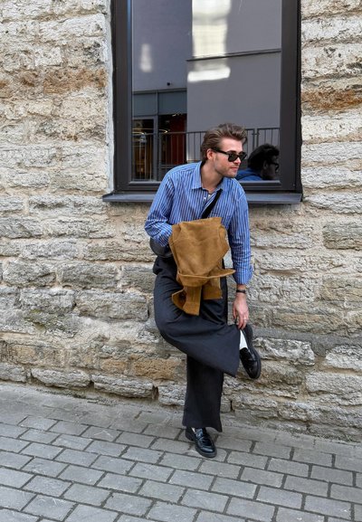 Hombre con gafas de sol ajusta su zapato negro mientras sostiene una chaqueta marrón, de pie junto a una pared de piedra bajo una ventana en una calle empedrada.