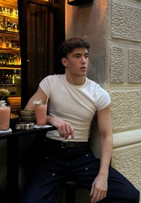 En ung mand i en tætsiddende hvid T-shirt og mørke jeans sidder afslappet ved et bord med lyserøde drikkevarer og snacks i en udendørs café.