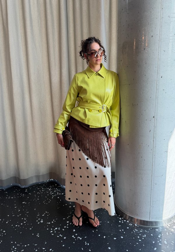 Femme portant une veste jaune ceinturée, jupe blanche à pois noirs avec franges marron, sandales noires et lunettes de soleil, debout près d’un pilier en béton.