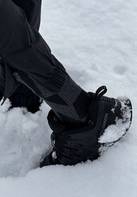 Chaussure de randonnée noire avec de la neige sur la semelle, portée avec une chaussette gris foncé et noire, debout dans une neige blanche profonde.