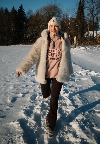 Mujer joven vestida con ropa de invierno, sonriendo y corriendo por un terreno cubierto de nieve con árboles al fondo.