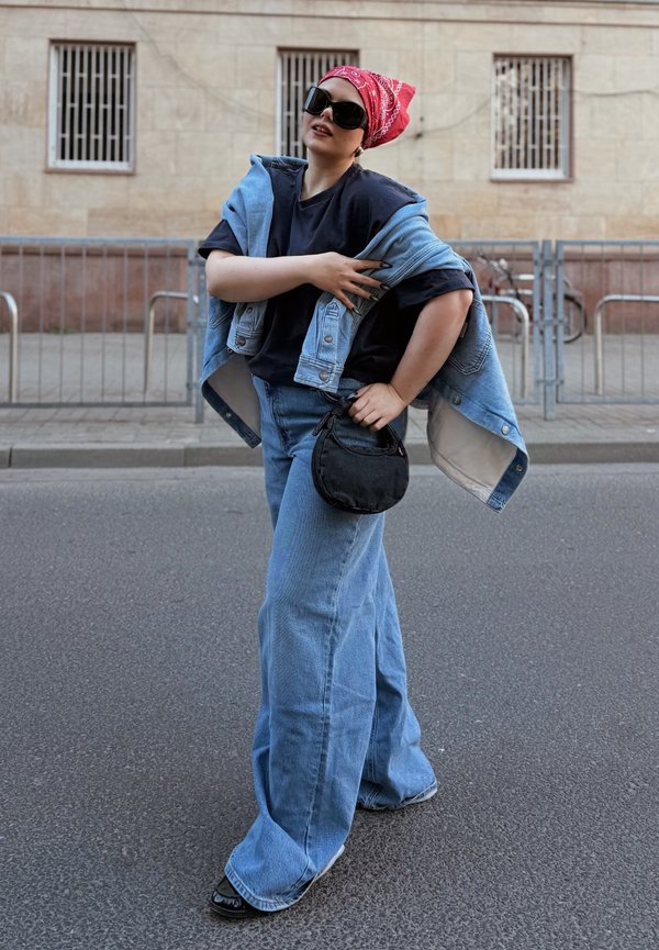 Person som bruker overdrevne blå jeans, svart skjorte, denimjakke over skuldrene, rød bandana, store solbriller, og holder en liten svart veske.