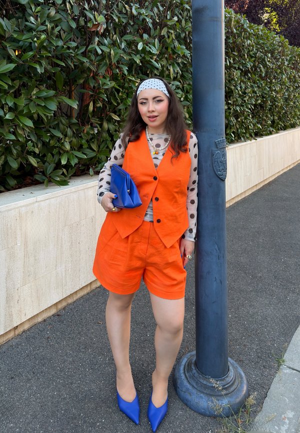 Kvinde i orange vest og shorts, prikket top, blå hæle og clutch, stående ved siden af en blå gadelampe på en brolagt fortov.