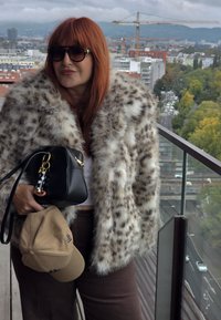 Leopard-print faux fur coat with a wide collar, paired with a black handbag featuring a gold keychain and a beige cap. Brown pants underneath.