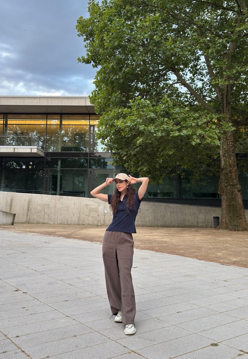 Woman wearing a beige cap, navy shirt, and brown pants stands on a paved area near a modern glass building and large green tree.