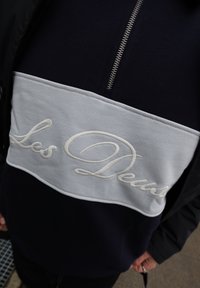 Άτομο που φοράει μπλε σκούρο και λευκό πουλόβερ με φερμουάρ στο μπροστινό μέρος και κεντημένο κείμενο "Les Deux" στο στήθος.