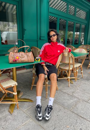 Mujer con chaqueta roja y pantalones cortos negros sentada en una cafetería al aire libre con café helado, bolso y cámara sobre una mesa verde.