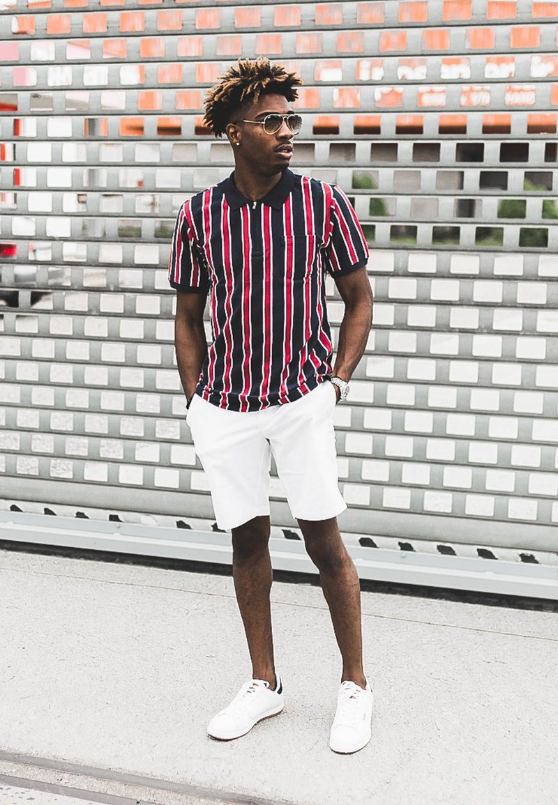 Rojo y azul marino a rayas, camisa de manga corta, pantalones cortos blancos, gafas de sol y zapatillas blancas. Fondo simple con un patrón geométrico.