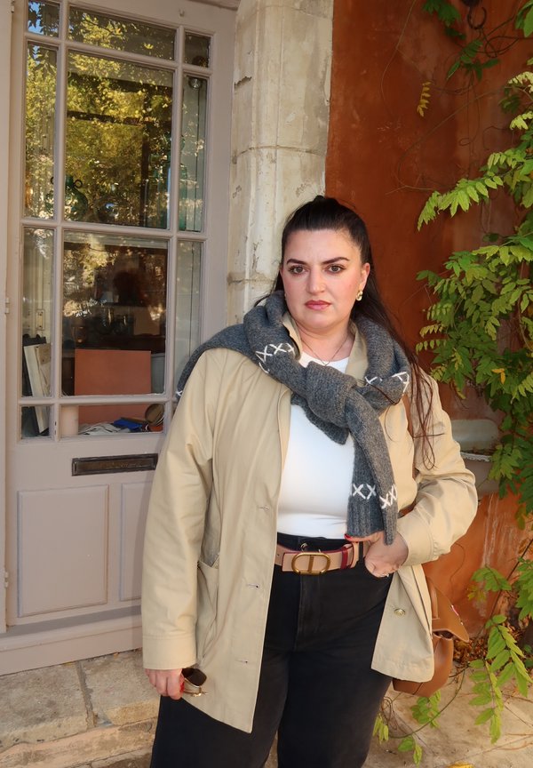 Beige Aussenjacke, weisses Oberteil, schwarze hochgeschlossene Hose, grauer Schal mit weisser Naht, goldene Ohrringe, hält Sonnenbrille, steht neben einer Tür.