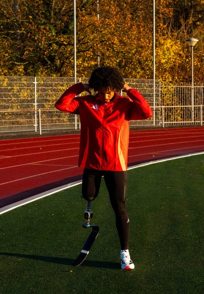 Atleta con prótesis de carrera ajusta su chaqueta roja en una pista al aire libre con árboles otoñales y una cerca de fondo.
