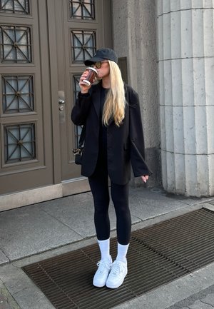 Blazer negro sobre una blusa negra, leggings negros y zapatillas blancas. Gorra de béisbol y taza de café. De pie en una acera cerca de una pared gris texturizada.