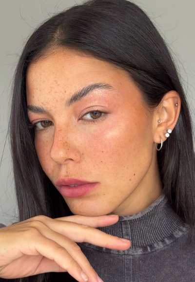 Mujer con cabello largo y oscuro y pecas, que lleva pendientes y un suéter gris de cuello alto acanalado, apoyando la barbilla en la mano sobre un fondo liso.