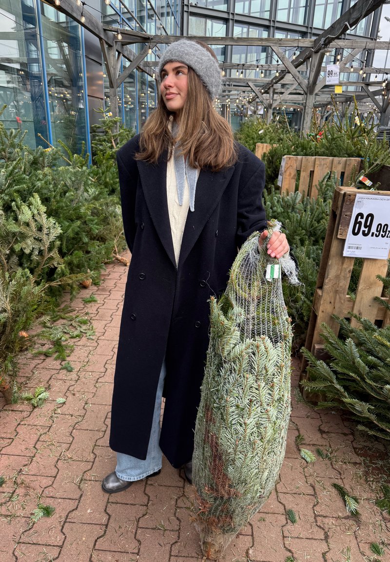 Une personne portant un manteau noir et un bonnet en tricot gris tient un sapin de Noël en filet, entourée de verdure et de structures d'exposition en bois.