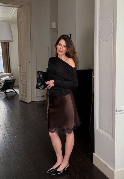 Mujer con blusa negra sin hombros, falda marrón con encaje y zapatos negros puntiagudos sosteniendo un bolso negro en un espacio de vida moderno.