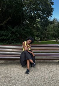 Osoba w jasnożółtej bezrękawnikowej bluzce i czarnej spódnicy siedzi na drewnianej ławce w parku, otoczona zielenią i ludźmi w tle.